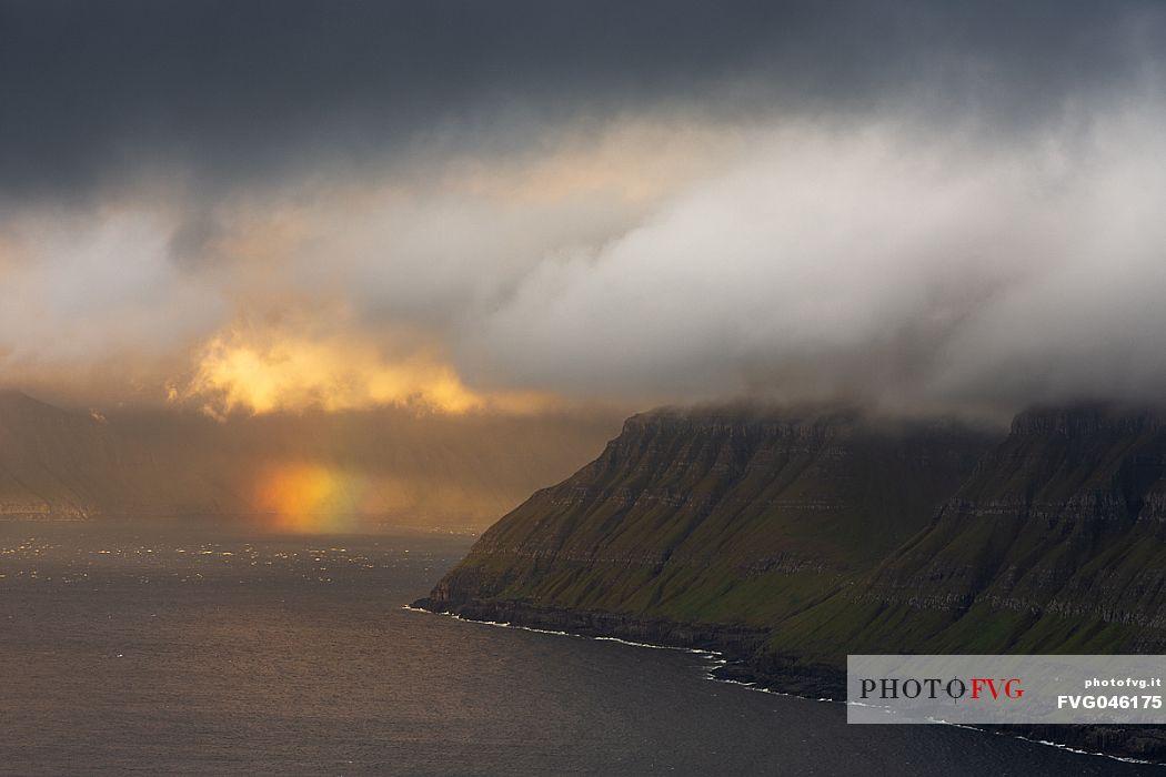 Beauttiful light and rainbow through the dense clouds of the coast, Faeroe islands, Denmark, Europe